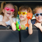 drei Kinder sitzen mit Sonnenbrille im Auto und lachen
